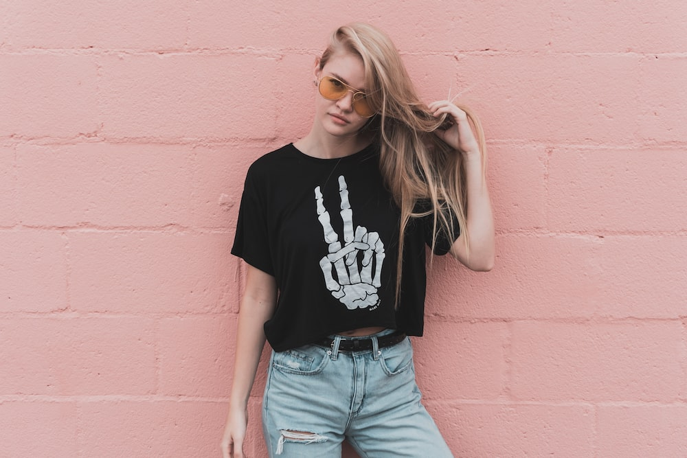 A Woman Wearing Shades, Jeans, and a Black Custom-Designed T-Shirt with a White Skeleton Hand Making a Peace Sign